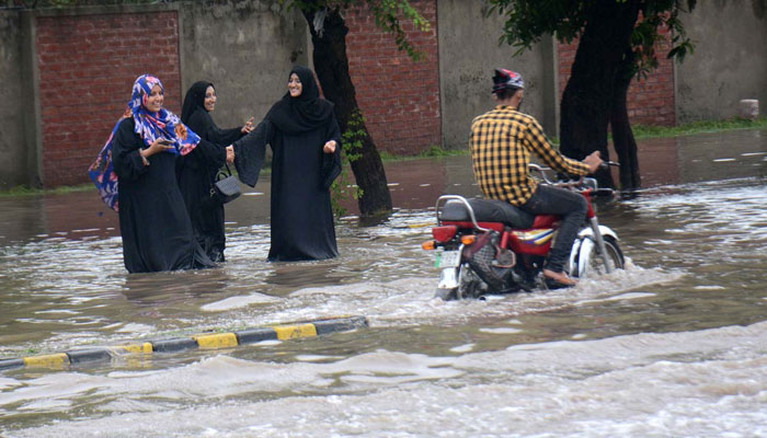A motorcyclist passing by three young women through accumulated rainwater on a road in Lahore on July 21, 2022. Photo: APP