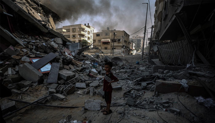 A child is seen standing among the debris of buildings as smoke is seen billowing in the background in Gaza. — X/UN