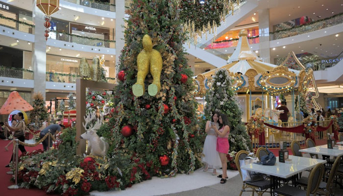 Shoppers taking photos stand beside a gag decoration depicting a reindeer crashing into a Christmas tree at a shopping mall in Kuala Lumpur on December 7, 2023. — AFP