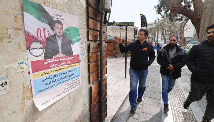 Men walk past a wall bearing an electoral campaign poster, ahead of the country’s upcoming parliamentary elections, in Tehran on Saturday. — AFP