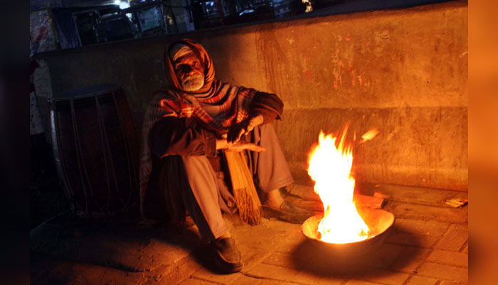 An aged man warms his hand on bonfire to protect himself from cold waves during the winter season, at Samanabad area in Lahore on December 19, 2024. — PPI