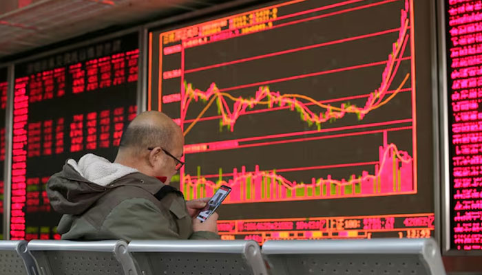An investor looks at his mobile phone in front of a board showing stock information at a brokerage office in Beijing, China January 2, 2020. — Reuters