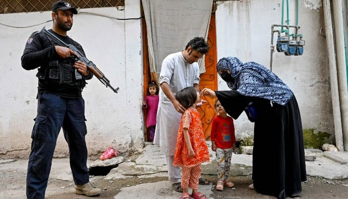 An elite police personnel (left) standing guard as a health worker (right) administers polio drops to a child during a door-to-door poliovirus vaccination campaign on the outskirts of Peshawar on October 29, 2024. —AFP