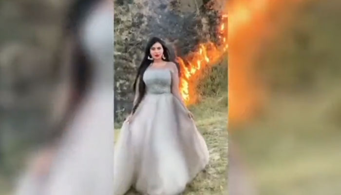 Woman posing for a photo as a fire rages in a field behind her. — Sscreengrab via Twitter/@rinasaeed