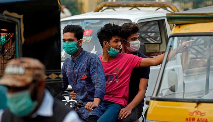ماسک پہنے تین نوجوان پاکستان کی ایک مصروف سڑک پر موٹر سائیکل پر سوار ہو رہے ہیں۔  — اے ایف پی/فائل