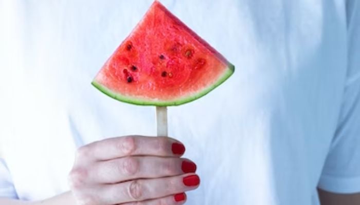 Image shows a person holding a slice of watermelon on a stick.— Unsplash