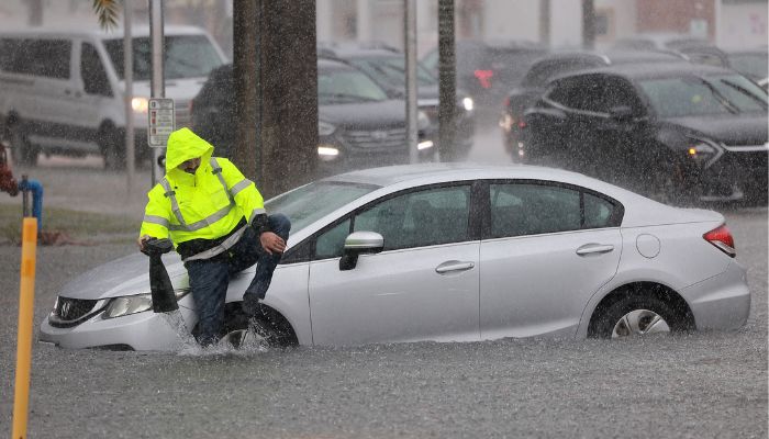 12 اپریل 2023 کو ڈینیا بیچ، فلوریڈا — اے ایف پی میں ایک ڈرائیور سیلاب زدہ سڑک پر کھڑی گاڑی کے ساتھ کھڑے اپنے جوتوں سے پانی نکال رہا ہے۔