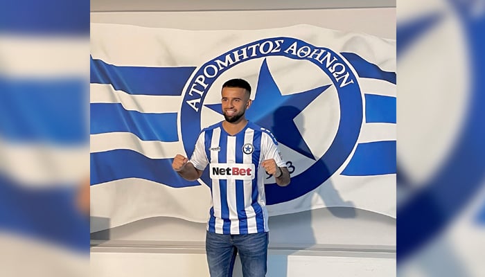 Adil Nabi poses in front of the flag of Atromitos Football Club. — Twitter/@NabiAdil