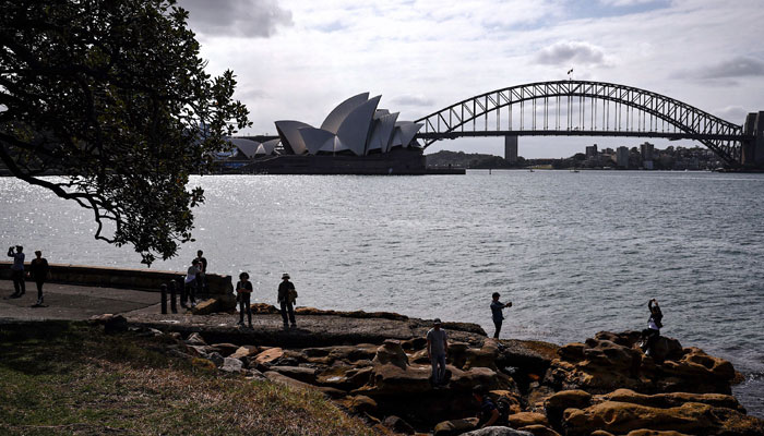 Tourists stand on rocks as they take photographs in front of the Sydney Opera House and Sydney Harbour Bridge on a spring day on November 13, 2023. — AFP