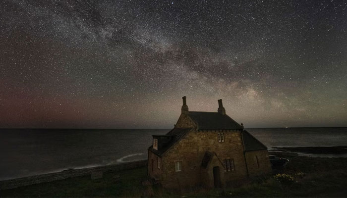 This image shows a cottage by the shore under a starry night sky. — The Independent via PA